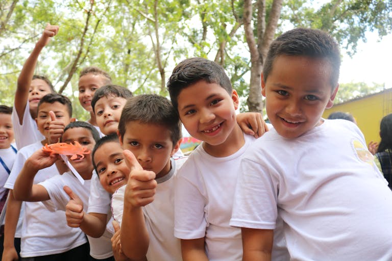 Group of happy children outdoors smiling and giving thumbs up, enjoying a sunny day.
